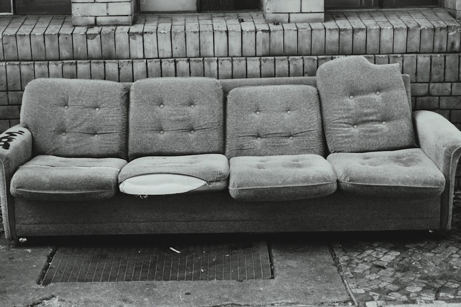 A three-seater fabric sofa in a faded gray colour is positioned outdoors against a brick wall. The sofa has a tufted backrest with visible buttons and a worn appearance, with one of the seat cushions missing, revealing a white fabric underneath. An armrest is visible on each side, with the left armrest showing a partially visible label or marking. The sofa is placed on a pavement surface, with a metal drainage grate directly in front of it. Behind the sofa, the brick wall consists of horizontally aligned, weathered bricks. The scene appears to be part of a home relocation or furniture disposal process, with Man with Van Cambridge Heath likely involved in the removal or transport of this item, fitting within the context of house removals and furniture transport.