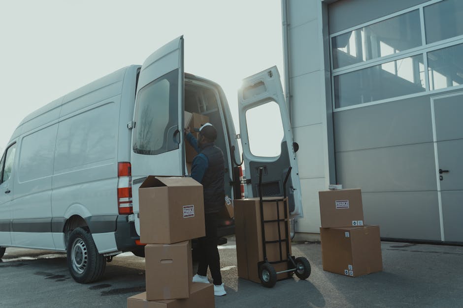A professional mover from Man with Van Cambridge Heath is loading a white moving van parked outside a modern commercial building with large glass windows during daylight. The mover, wearing a dark ves