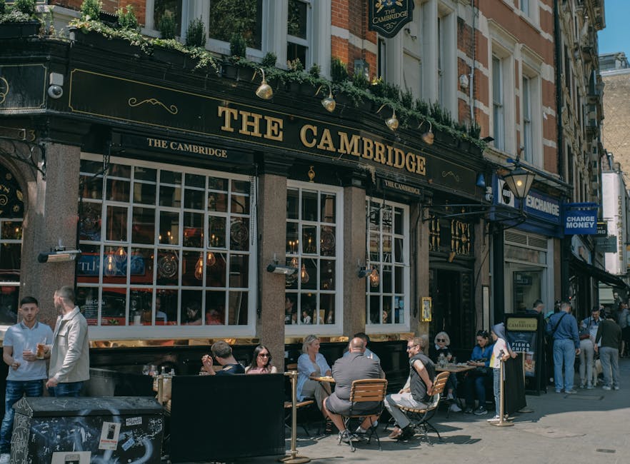 The image shows the exterior of a traditional pub named 'The Cambridge,' located on a busy street during daytime, with large multi-pane windows and a black and gold sign above the entrance. Several people are sitting at outdoor tables, some engaged in conversation, while others are enjoying drinks. The tables and chairs are made of wood and metal, placed directly on the pavement in front of the pub. A few pedestrians are walking along the street, and others are gathered near the entrance, possibly waiting to enter or chat. The pub's façade features decorative lighting fixtures and greenery on the upper ledge. This scene likely depicts a lively urban area suitable for home relocation planning or furniture transport, as part of a professional removals process by [COMPANY_NAME], with logistics involving packing, loading, and transportation of household items, set against the backdrop of Cambridge Heath Road.
