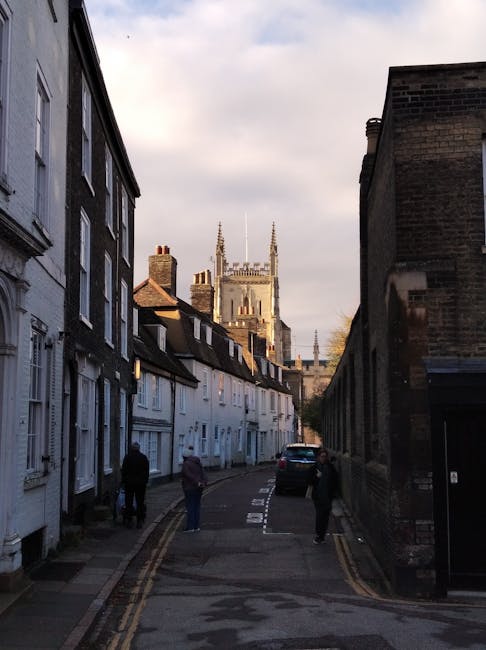 A narrow residential street on Cambridge Heath Road with white terraced houses on the left and a tall, dark brick building on the right. Several pedestrians are walking along the pavement, and a black van is parked near the end of the street. In the background, a large church or cathedral with tall spires is visible against a cloudy sky. This urban scene captures the typical setting for house removals or moving services, with close-proximity buildings and an active environment suitable for furniture transport and loading processes that a company like Man with Van Cambridge Heath might handle during home relocation projects.