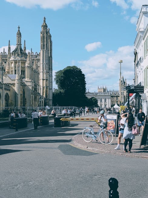 The image shows the exterior of a traditional pub named 'The Cambridge,' located on a busy street during daytime, with large multi-pane windows and a black and gold sign above the entrance. Several people are sitting at outdoor tables, some engaged in conversation, while others are enjoying drinks. The tables and chairs are made of wood and metal, placed directly on the pavement in front of the pub. A few pedestrians are walking along the street, and others are gathered near the entrance, possibly waiting to enter or chat. The pub's façade features decorative lighting fixtures and greenery on the upper ledge. This scene likely depicts a lively urban area suitable for home relocation planning or furniture transport, as part of a professional removals process by [COMPANY_NAME], with logistics involving packing, loading, and transportation of household items, set against the backdrop of Cambridge Heath Road.