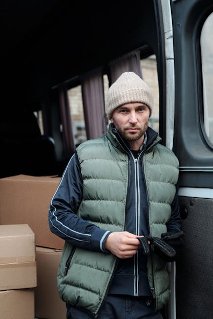 A man wearing a white t-shirt, blue jeans, and a black cap is sitting on the pavement beside the open rear door of a large white van during a house relocation. Inside the van, there are numerous brown