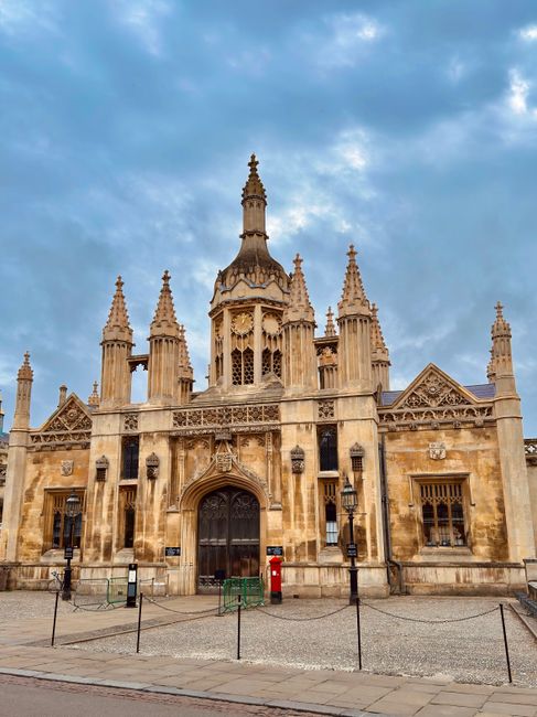 The image features an ornate, historical building constructed from light-colored stone with intricate Gothic architectural detailing, including numerous spires and decorative elements, set against a partly cloudy sky. In front of the building, there is a paved area with black and white bollards, some with horizontal stripes, and metal barriers. A red post box is visible near the entrance, which is flanked by two black lantern-style street lamps. The scene appears to be taken during daytime with natural light illuminating the facade. Although no furniture or moving equipment is visible in this outdoor setting, the image depicts a well-preserved landmark, possibly within an urban environment commonly associated with house removals or relocation services such as those offered by Man with Van Cambridge Heath. This setting can relate to the context of home relocation logistics, especially when planning moves near historic or notable buildings.