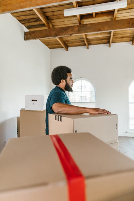 A man with dark skin and curly hair wearing a teal T-shirt is positioned indoors, standing behind a stack of large cardboard moving boxes during a home relocation process. The boxes are sealed with re