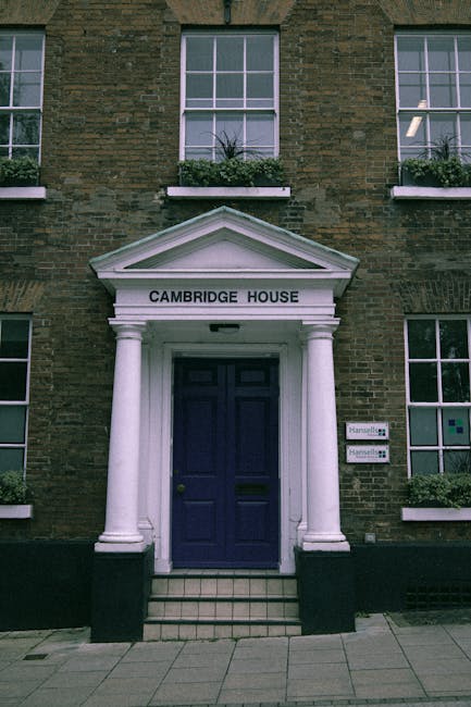 The image shows the front entrance of a brick building labeled 'Cambridge House' with a white triangular pediment supported by two white columns, above a dark blue door. The door is set within a small porch area with a tiled step leading up to it. There are multiple windows with white frames on the building front, some with flower boxes containing green plants. Outside, on the pavement, a black lifting trolley is visible, loaded with several cardboard boxes and wrapped furniture, indicating a home relocation process. The setting suggests a professional removals scene, typical of house moving or furniture transport services in Cambridge Heath, with Man with Van Cambridge Heath potentially operating in this context.