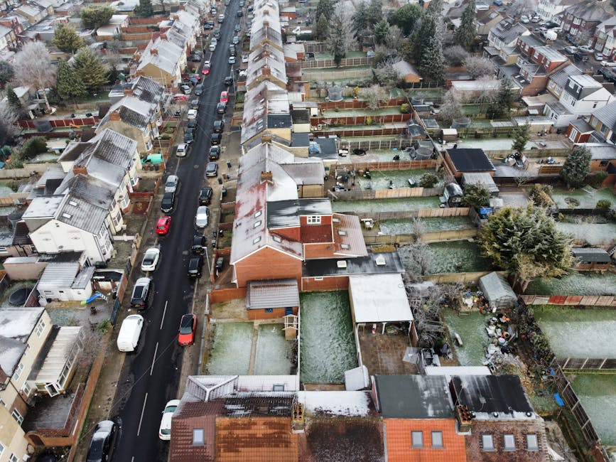 An aerial view of a residential street in Cambridge Heath showing parked cars lining both sides of a narrow road with snow-dusted rooftops and garden fences. The houses are mostly terraced with pitched roofs, some covered in snow, and small backyards enclosed by wooden fences. Several gardens contain garden furniture, sheds, and lawn areas, with some showing garden waste or clutter. A few houses have external chimneys, and the scene is captured during daylight under overcast conditions, creating a neutral, natural setting typical for home relocation and furniture transport activities. The image reflects a typical neighbourhood with consistent row houses and closely packed gardens, relevant to the context of house removals and moving logistics in the area, with occasional mention of companies like Man with Van Cambridge Heath involved in packing and moving services.