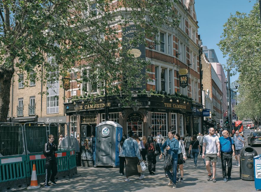 A busy street scene outside a traditional pub building called 'The Cambridge' located on the corner of a street in Cambridge Heath. The building has a dark exterior with signage displaying the pub's name and decorative hanging lanterns. Several pedestrians are walking along the pavement, some carrying shopping bags or backpacks, and dressed in casual attire suited for a warm day. In the foreground, a parcel box and portable toilets are positioned near a green temporary fence, indicating ongoing setup or work related to the area. A man is seen lifting or moving items, possibly furniture or packing materials, onto a van parked nearby, with the van's rear doors open. The street is lined with trees providing partial shade, and other commercial buildings are visible further down the street, along with parked cars and a red double-decker bus in the distance. This scene illustrates a typical urban environment where house removals or furniture transport processes might take place, supported by local businesses such as Man with Van Cambridge Heath engaged in home relocation services.