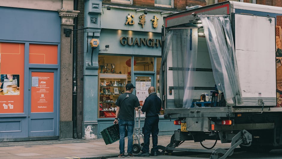 Two men from Man with Van Cambridge Heath are engaged in the home relocation process outside a storefront with Chinese signage in Cambridge Heath. One man, dressed in dark clothing, is carrying a black plastic crate, while the other, wearing a cap and a black jacket, stands nearby with a small hand truck ready to load or unload furniture or boxes. A large delivery van with an open side door is parked on the pavement, revealing the interior partially covered with clear plastic sheeting for protection during furniture transport and packing and moving activities. The building's facade features decorative molding and large windows, with a visible retail display inside. The scene is illuminated by daylight, emphasizing the organized approach of the moving process as part of a professional removals service, subtly illustrating the logistics involved in house or office moves, including furniture handling and packing materials, with a focus on outdoor loading and unloading operations.