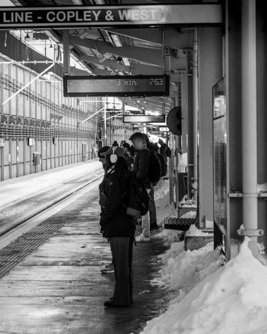 Black and white photograph of a group of people waiting on a snow-covered platform at Cambridge Heath station, part of the Copley & West line. The individuals are bundled in winter clothing, with some carrying backpacks and bags, and they stand near a shelter with a bench. Overhead, an electronic display shows the next train departure details, including the time and destination. The platform features tactile paving along the edge for safety. To the right, a large snow pile partially covers the lower part of the building structure, indicating recent snowfall. In the background, there are metal railings, station signage, and tracks extending into the distance. The scene portrays an everyday moment of waiting for train transport, which relates to the process of home relocation or moving logistics when planning house moves in snowy conditions. This image is associated with Man with Van Cambridge Heath, specialising in removals and moving services, illustrating the importance of punctuality and safety during transportation of belongings in winter.
