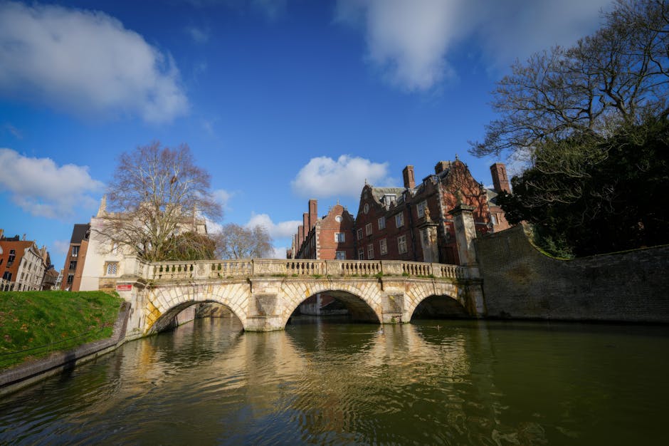 A picturesque daytime view of a historic stone bridge with three arches spanning over a calm river, with clear reflections visible on the water surface. The bridge has a decorative stone balustrade along its length, and a small red and white sign is attached to the side. In the background, there are several multi-storey red brick buildings with chimneys, characteristic of traditional architecture, along with leafless trees indicating a colder season. The sky is bright blue with scattered white clouds, and sunlight illuminates the scene, highlighting the textures of the stonework and the surrounding environment. The scene captures a tranquil moment in an urban area, suitable for house removals or relocation services, with a focus on the external environment just before a home relocation or furniture transport activity. This image is relevant for companies like Man with Van Cambridge Heath that specialize in removals and moving logistics services, providing a visual context for city-based home moves or furniture transport projects.